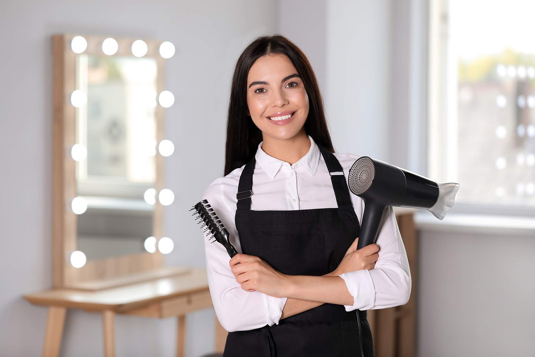 A smiling hairstylist holding a hairdryer and brush inside a modern hair salon